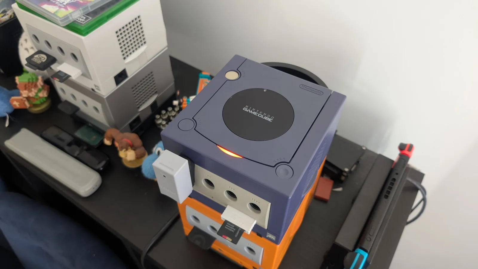 Indigo GameCube, powered on with its red LED lit, stacked on an orange GameCube, a pearl white GameCube, amiibo and a Switch around it, at fleet standard after the retrobrite.