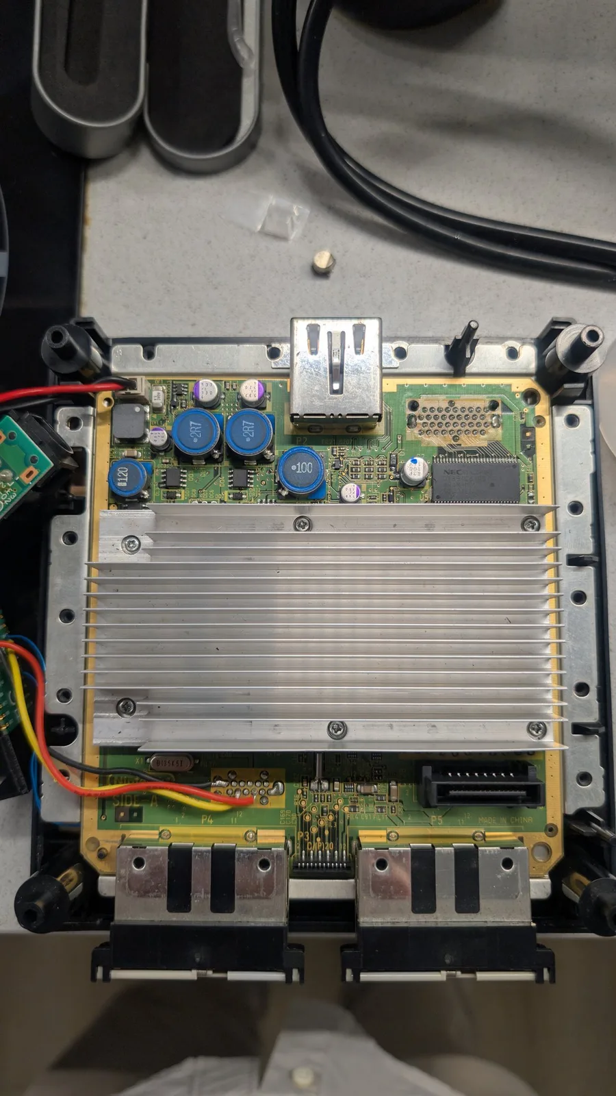 The same PAL DOL-101 GameCube mainboard with the PicoBoot mod installed, its blue inductors and aluminium heatsink visible; the recap done, a new capacitor marked with a blue dot now sits where the black-striped cap had been, and the old cap is visible on the counter behind the console.