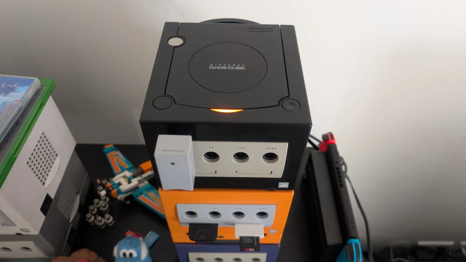 A black Nintendo GameCube powered on and being tested after its recap (this post's console), a WaveBird wireless receiver plugged in. It is stacked on a spice-orange GameCube that has a white SD storage interface connected, above a dark blue/indigo GameCube, with a docked Nintendo Switch alongside.