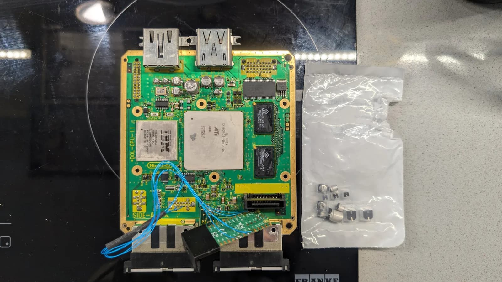 GameCube DOL-CPU-11 mainboard with the IBM Gekko CPU and the ATI/NEC Flipper chip (bare metal top, no heatsink fitted), the proprietary AV/digital output ports at the top edge (not USB), the RAM chips and blue kynar PicoBoot wires at the bottom edge, on a black surface beside a Console5 capacitor-kit bag.