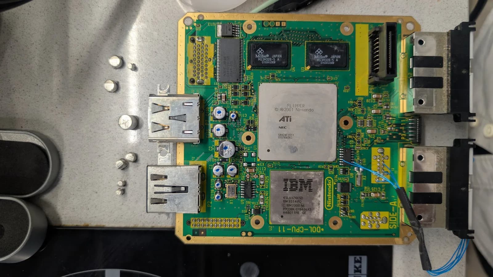 GameCube DOL-CPU-11 mainboard showing the ATI/NEC Flipper chip and the IBM Gekko processor, its newly replaced capacitors dotted with blue felt pen to mark them done, with blue kynar PicoBoot wires at the lower-right corner.