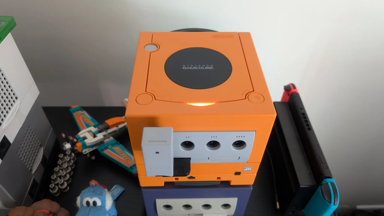 Spice orange GameCube, lid closed and powered on (orange LED lit), with a memory card in the slot and a WaveBird wireless receiver in controller port 1, stacked on top of an indigo GameCube; a Nintendo Switch and a white console sit alongside.