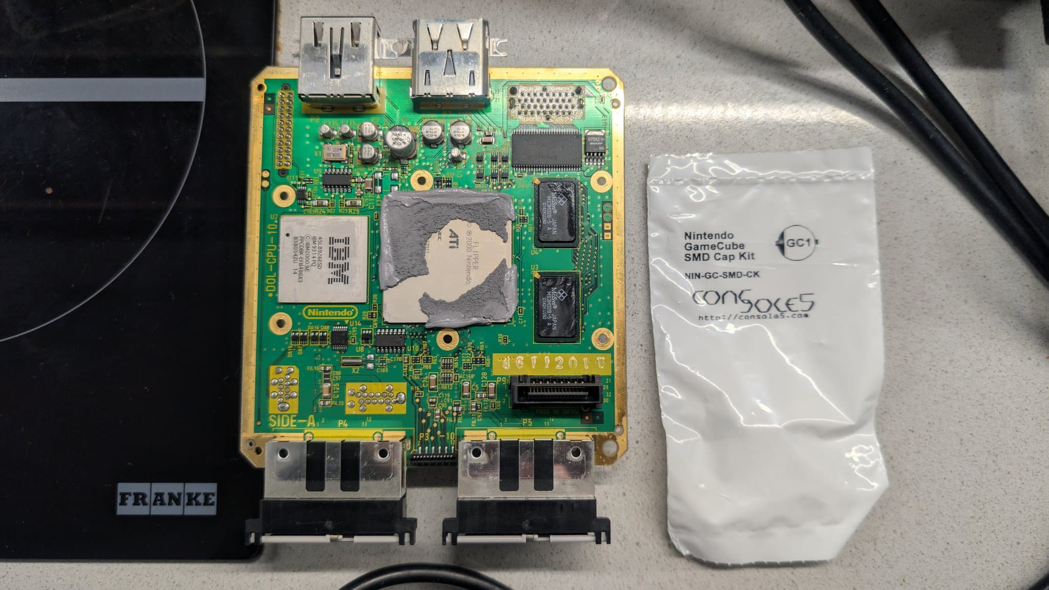 Nintendo GameCube mainboard (side A) with exposed CPU and RAM chips, two HDMI ports, and SMD cap kit package during recap restoration project.