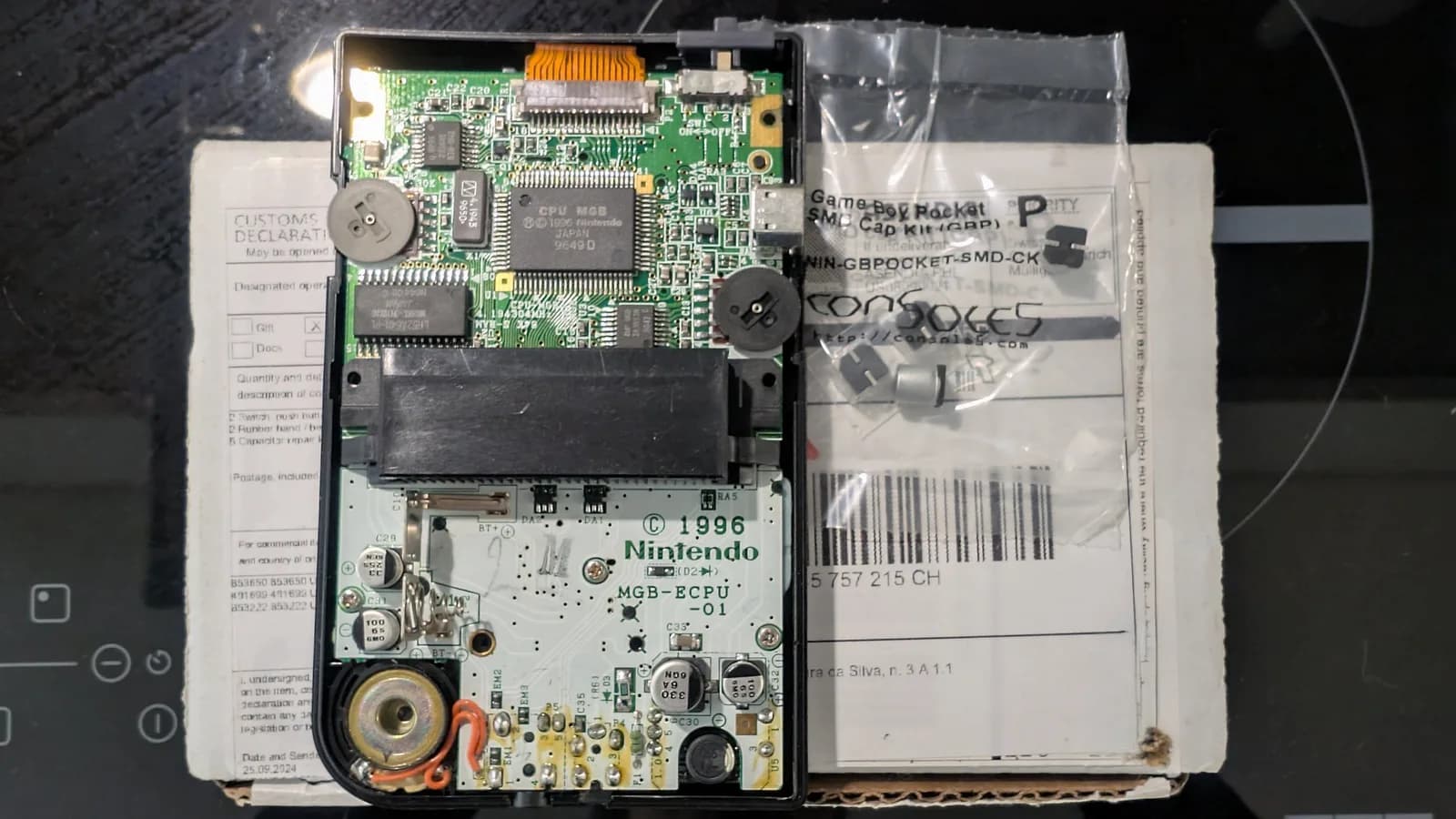 Black Game Boy Pocket motherboard in its shell, still fully populated: the CPU MGB chip, RAM, electrolytic capacitors, the two brightness and volume trimmer wheels, the cartridge connector and speaker. It rests on the Console5 box the cap kit shipped in (customs label showing), with the Console5 Game Boy Pocket SMD cap kit bag beside it.