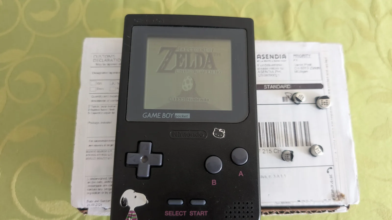 Reassembled black Game Boy Pocket powered on, showing the Zelda: Link's Awakening title screen, with a Hello Kitty sticker beside the embossed Nintendo logo, a Snoopy sticker on the lower front, and pink A/B/SELECT/START labels. It rests on the Console5 box the cap kit shipped in (customs label showing), the removed old capacitors sitting on it.