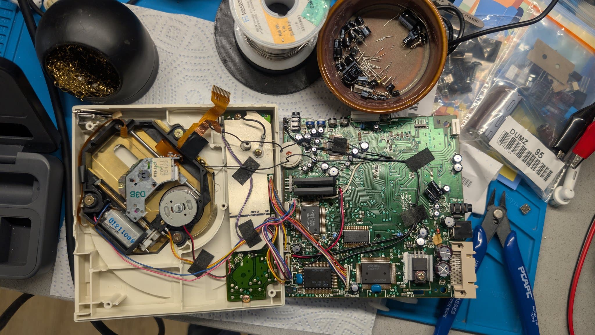 Disassembled PC Engine CD-ROM2 showing the now-recapped mainboard with fresh capacitors, the laser assembly, motor components and repair tools, with the old removed capacitors collected in a bowl at the top of the shot above the recapped board.