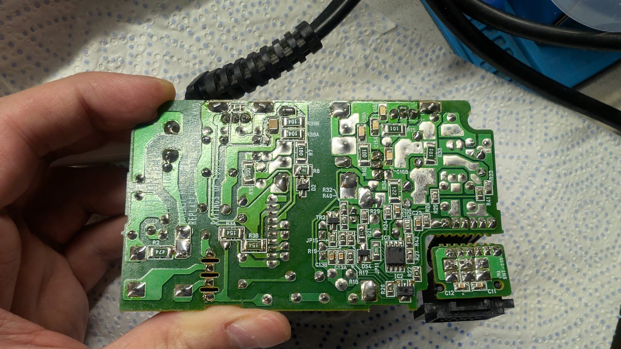Solder side of the Nintendo 64 power supply PCB held in hand, showing the reflowed solder joints near the power connector and wire terminals (the new capacitors are on the component side).