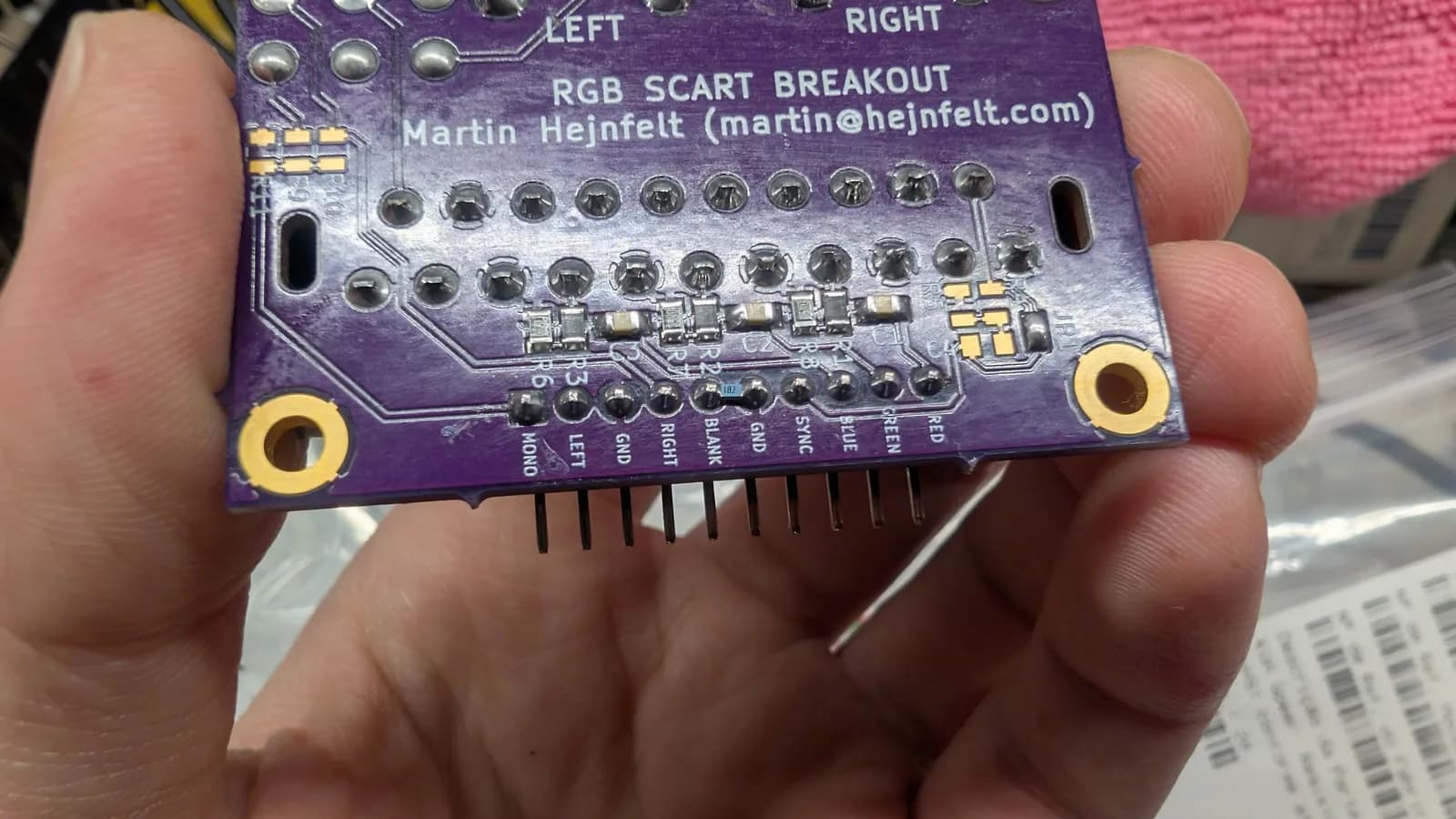 Purple RGB SCART breakout PCB by Martin Hejnfelt held in hand, showing solder joints, resistors, and labeled pin headers for LEFT/RIGHT audio and video signals.