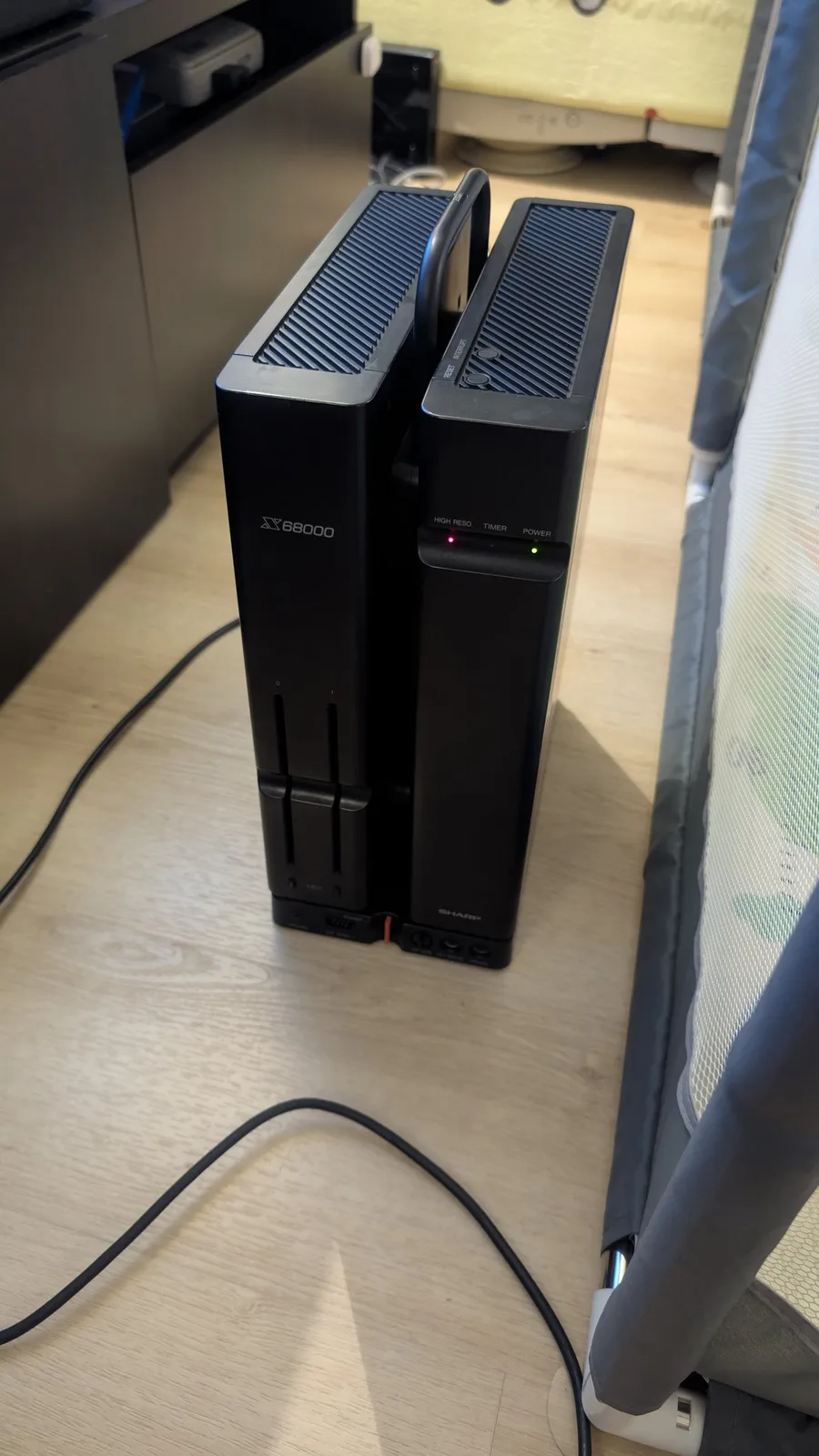 The Sharp X68000 on a wooden surface during the powered-on test with the VGA adapter and OSSC; its iconic design is two interlinked towers, the left one labelled X68000 and the right one showing the power LED (green) and the high-reso LED (red).