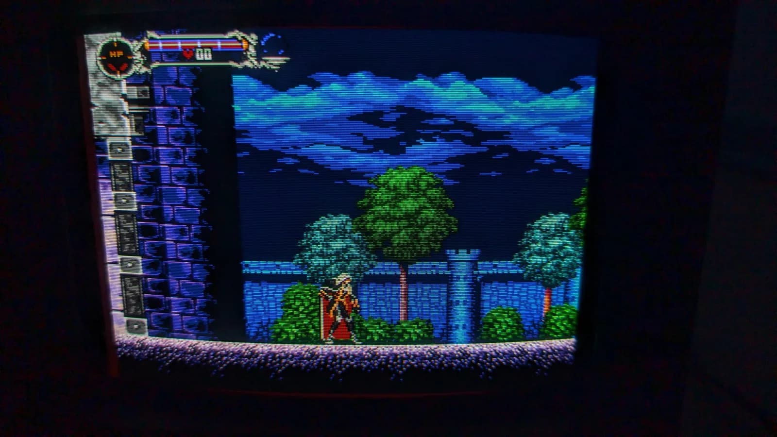 Sony PVM-9045QD running the Mega Drive homebrew demake of Castlevania: Symphony of the Night after the deflection-board recap, Alucard before the castle wall under a blue cloudy sky with trees, the HP bar and item column at the left.