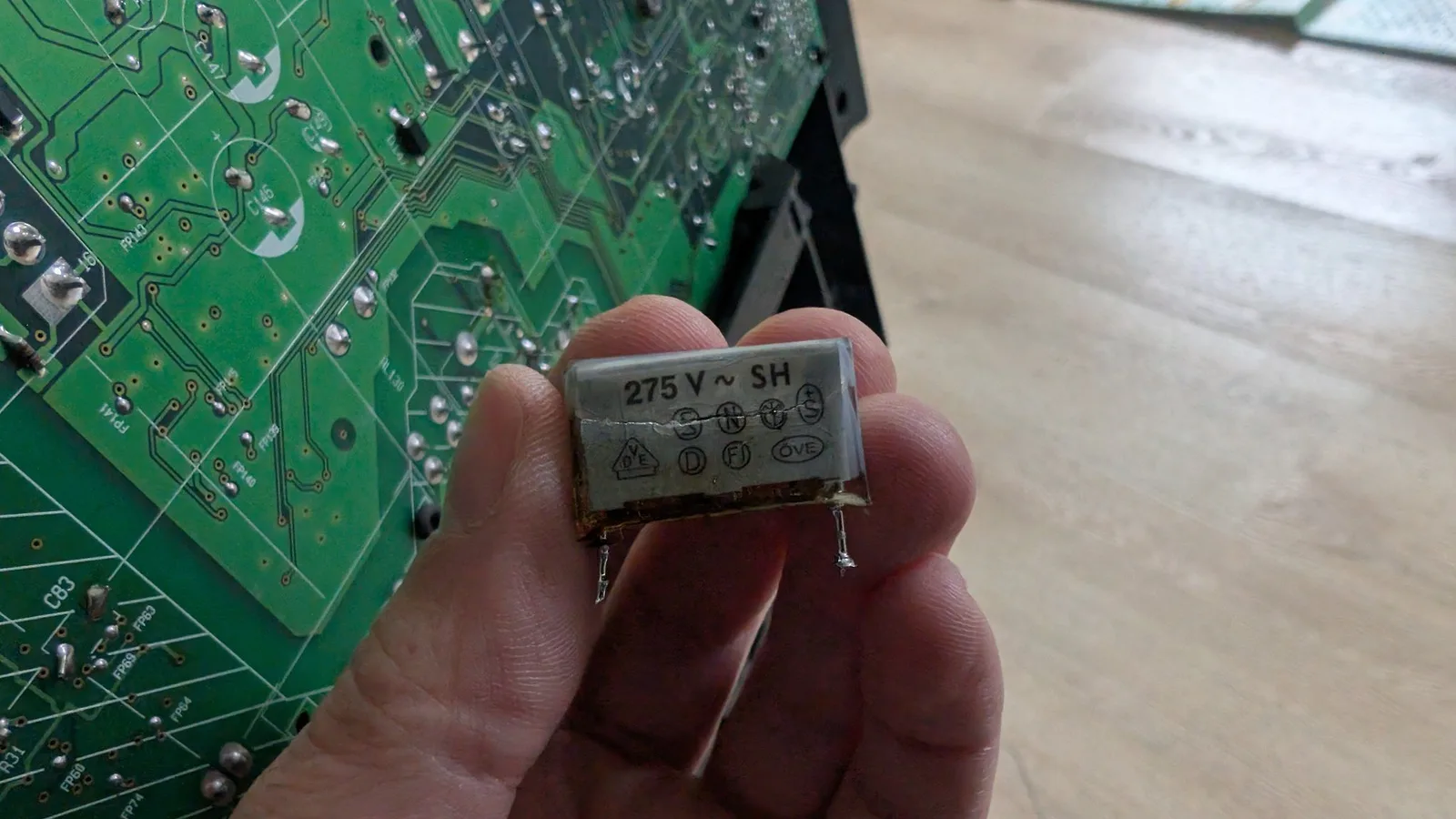 Damaged X2 safety capacitor labeled "275 V ~ SH" held in fingers, showing catastrophic failure with exposed internal metal components.