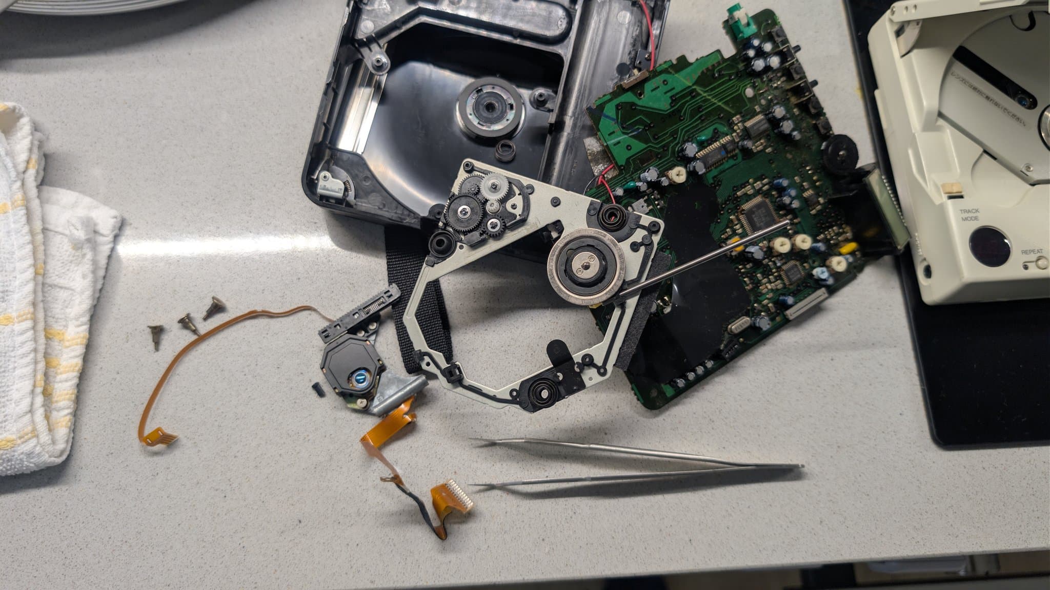 Disassembled Sony Discman, the donor unit the replacement laser pickup was harvested from, showing its laser mechanism, circuit boards and gears with tweezers on the work surface.