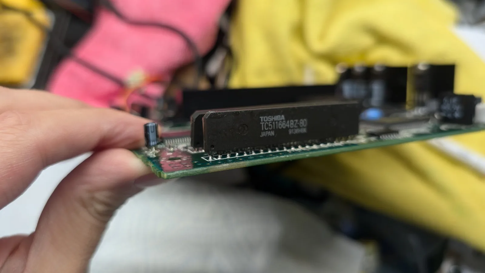 Hand holding Toshiba TC511664BZ-80 RAM chip over green circuit board during Wondermega repair installation.
