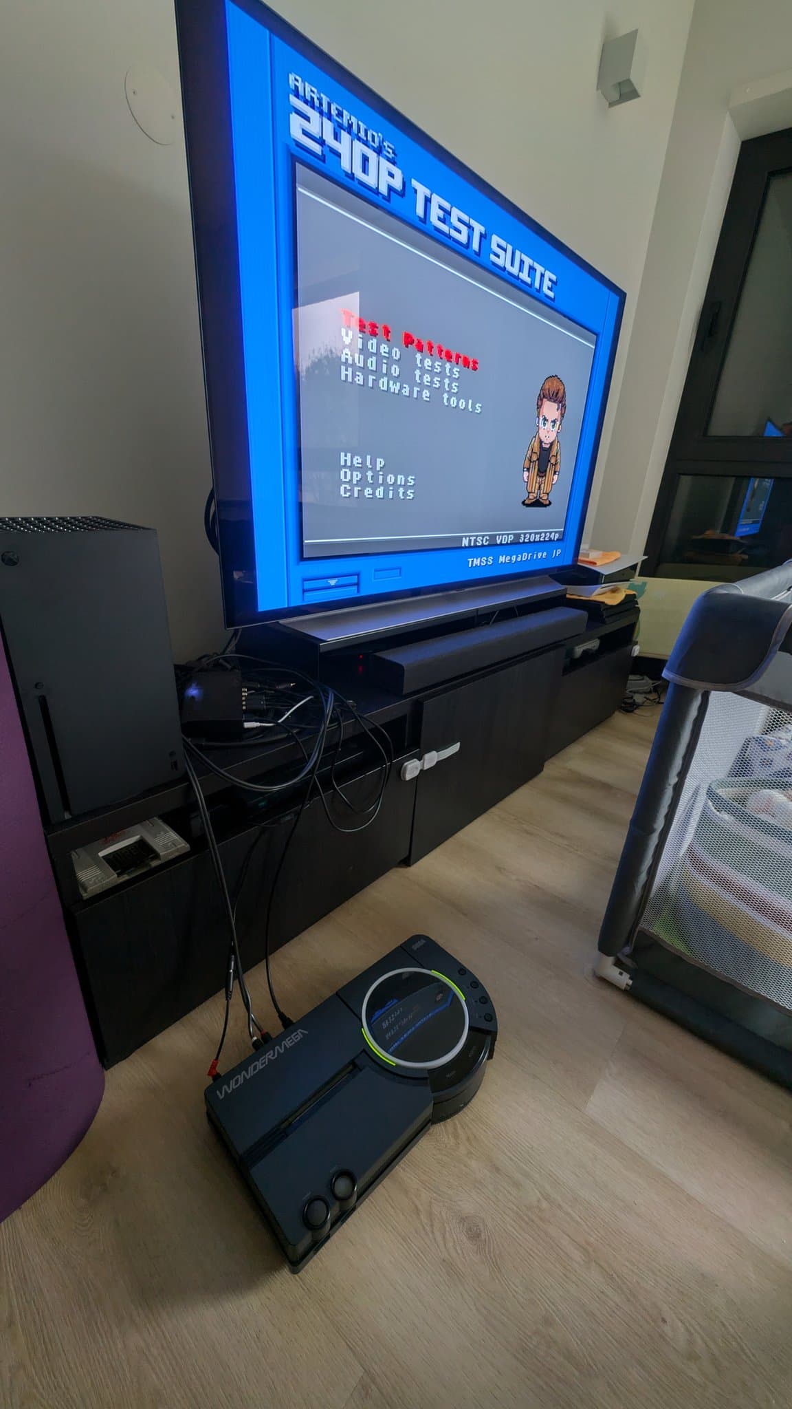 Sega Wondermega connected to TV displaying 240p test suite menu via flash kit programmer; controller visible on floor.