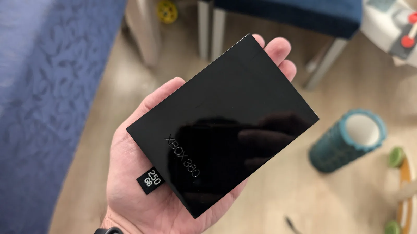 Hand holding a glossy black Xbox 360 hard drive caddy embossed 'XBOX 360' with a 250 GB label, over a wood floor with children's toys in the background.