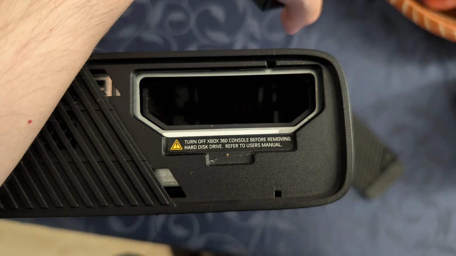 Close-up of the Xbox 360 E's empty hard drive bay with the cover off, showing the yellow-triangle warning label to turn off the console before removing the hard disk drive.