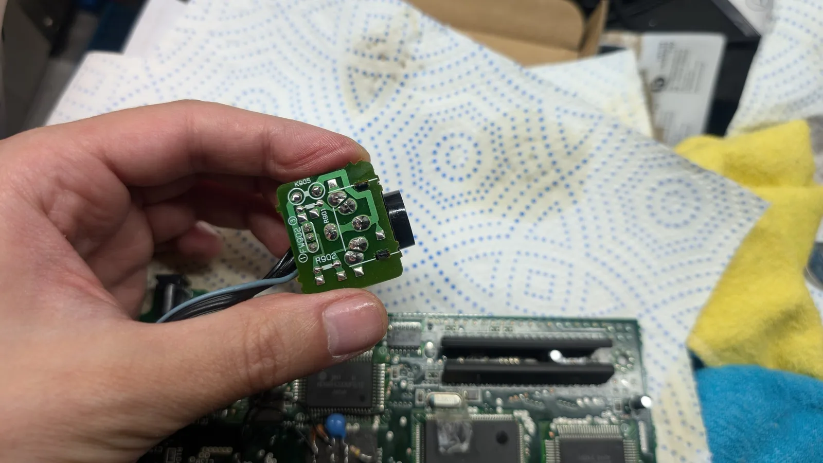 Hand holding recapped green AV board with black connector; solder joints reflowed during Wondermega repair work.