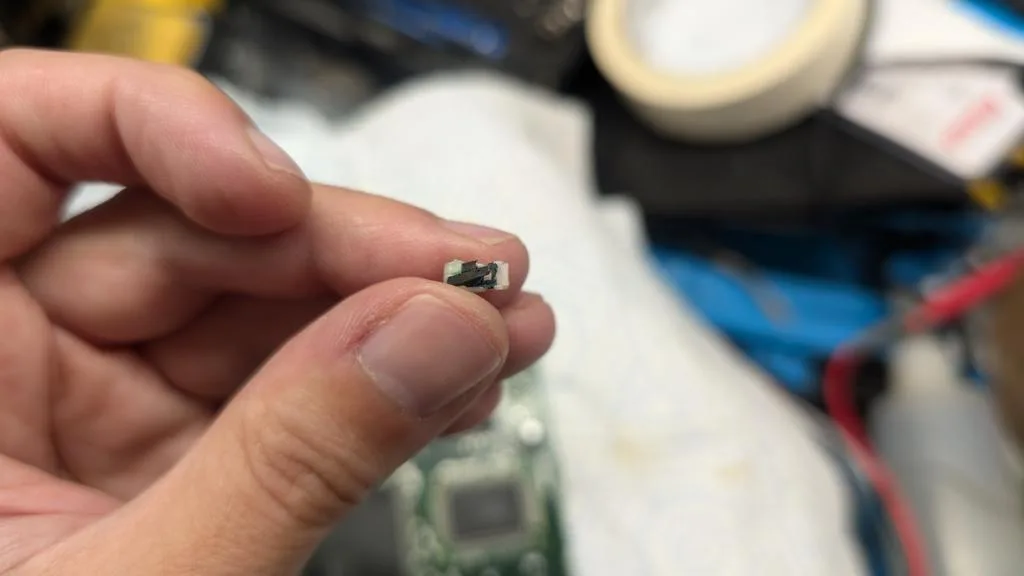 Corroded cartridge detection button held in tweezers over workbench with tape and repair supplies visible.