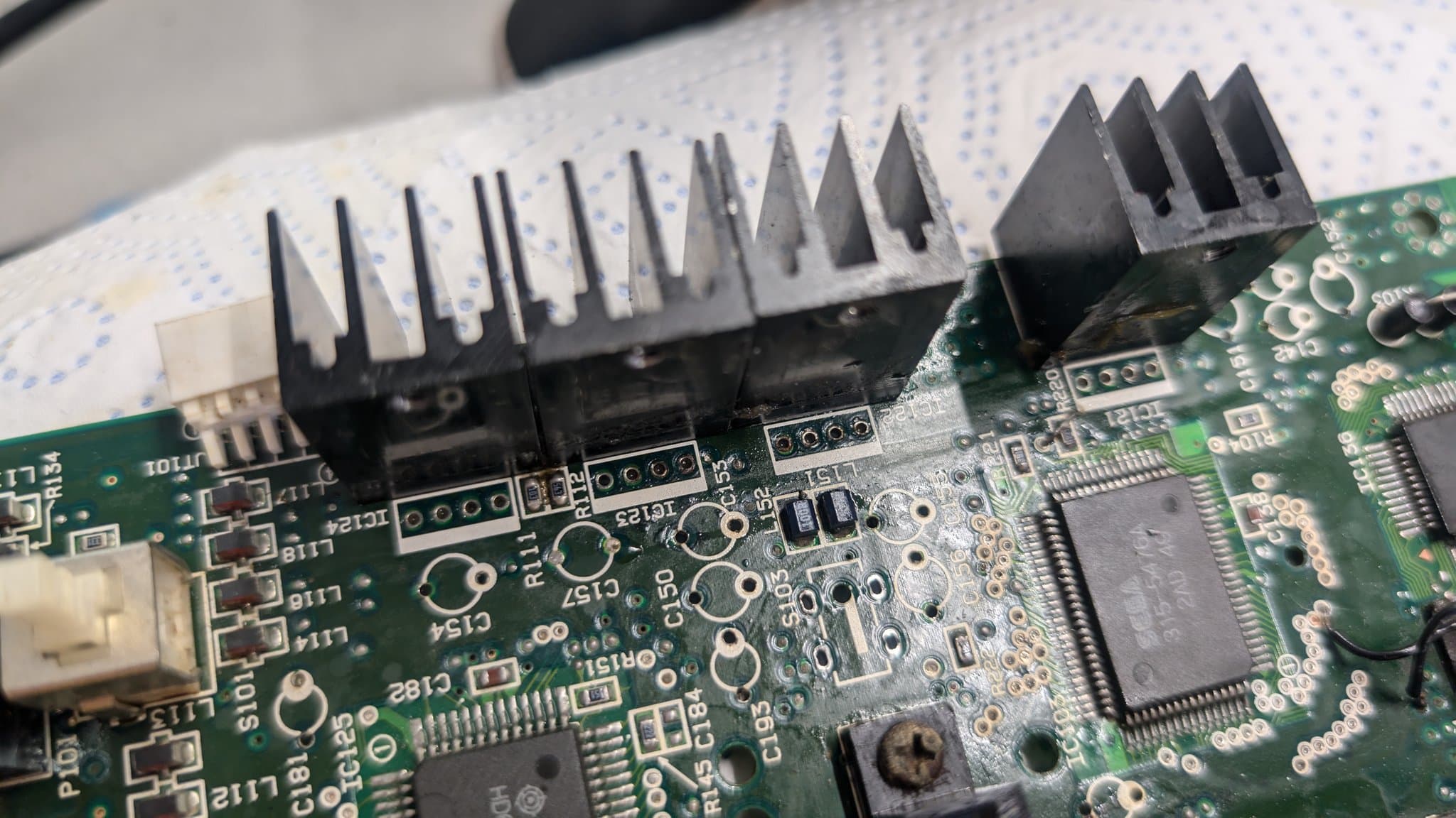 Sega Wondermega PCB with multiple solder joints being reflowed using flux; black heatsinks visible on ICs during corrosion and electrolyte cleanup.