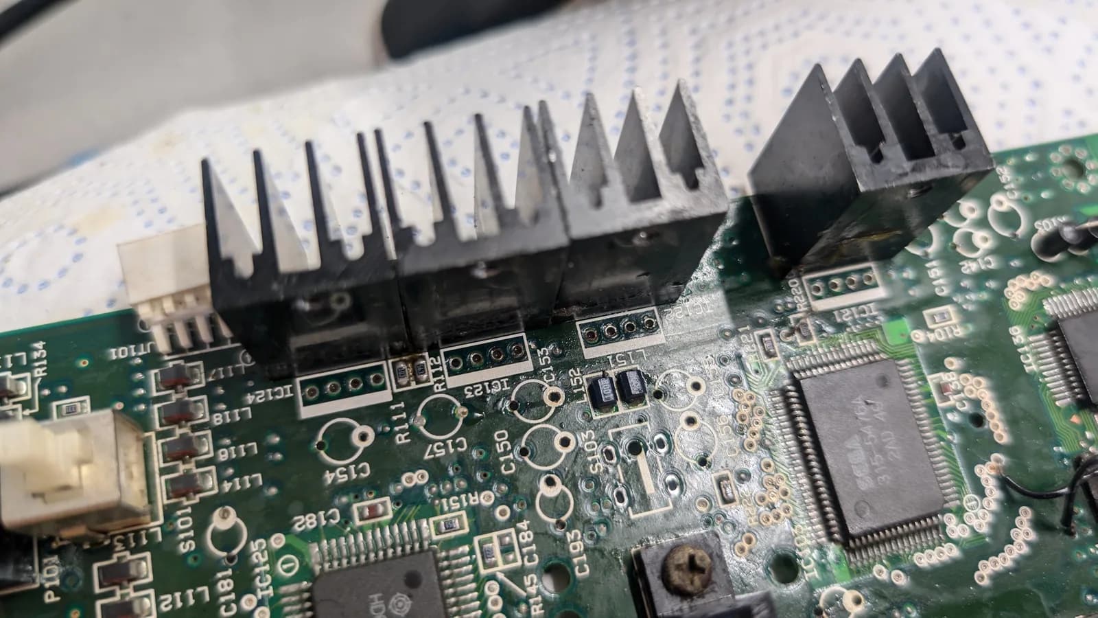 Sega Wondermega PCB with multiple solder joints being reflowed using flux; black heatsinks visible on ICs during corrosion and electrolyte cleanup.