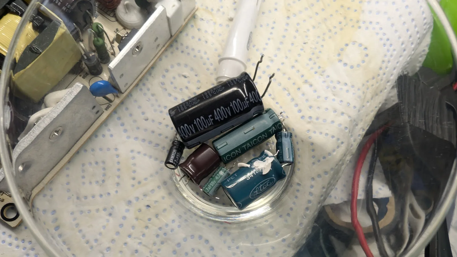 Old bargain-bin capacitors removed from the Xbox 360 E power brick, sitting in a clear glass bowl on a paper towel: a black 100µF 400V cap, a teal Taicon cap, a teal Ltec cap, green Samxon caps, and a brown capacitor.