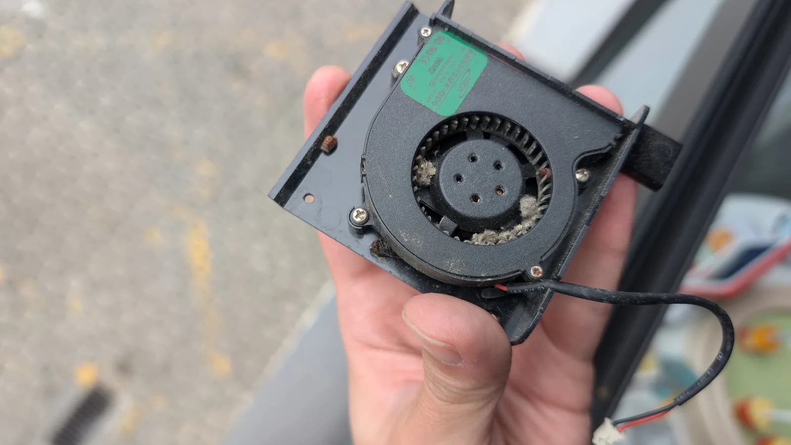 The Xbox 360 E power brick's centrifugal blower fan held in hand, its intake and blades clogged with heavy dust, lint and bug carcasses (with more beneath it), a green model-number label on the fan housing.