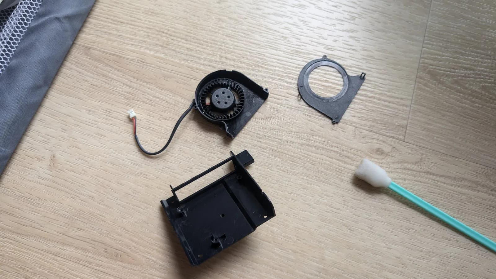 Disassembled Xbox 360 E power-brick parts on a wood floor: the cleaned black centrifugal blower fan with its connector and wire, the fan's shroud ring, a black plastic housing, and a green-handled foam cleaning swab.