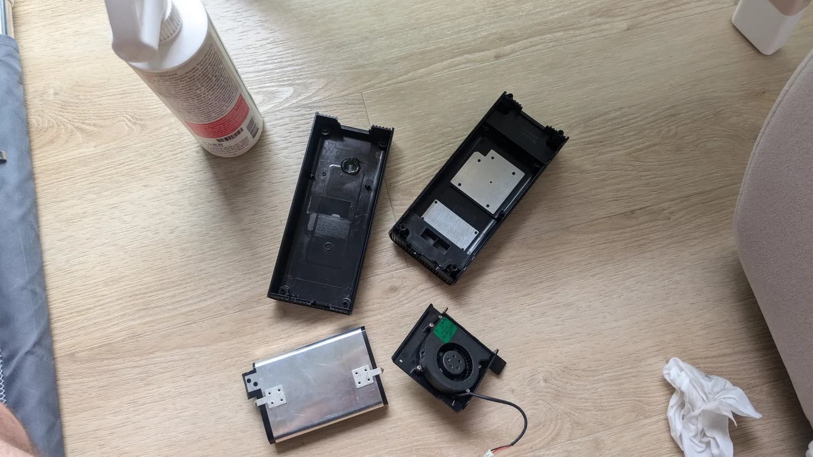 Disassembled Xbox 360 E power brick on a wood floor: the two halves of the black plastic housing, a metal heatsink plate, and the black blower fan, beside a bottle of isopropyl alcohol and a tissue.
