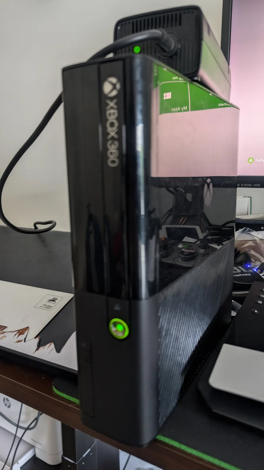 Restored Xbox 360 E console with its green power ring lit, in front of a monitor showing the dashboard, the PSU brick behind it with its green indicator also lit.