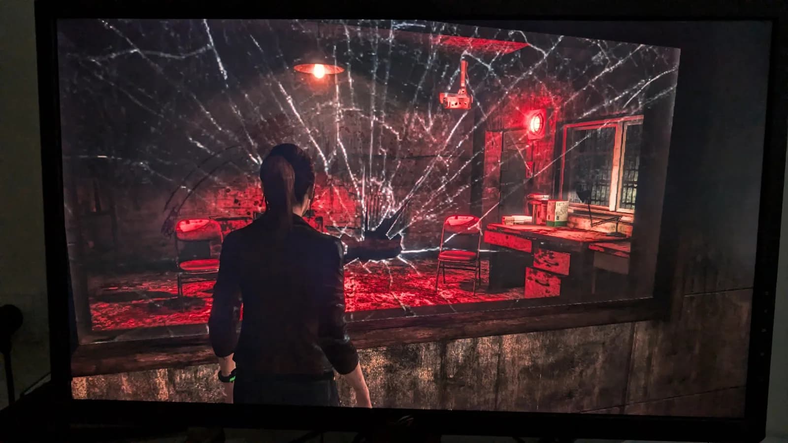 A monitor showing Resident Evil Revelations running on the Xbox 360 E after its PSU recap: a third-person female character in a dark, red-lit interior room, viewed through a shattered pane of glass.