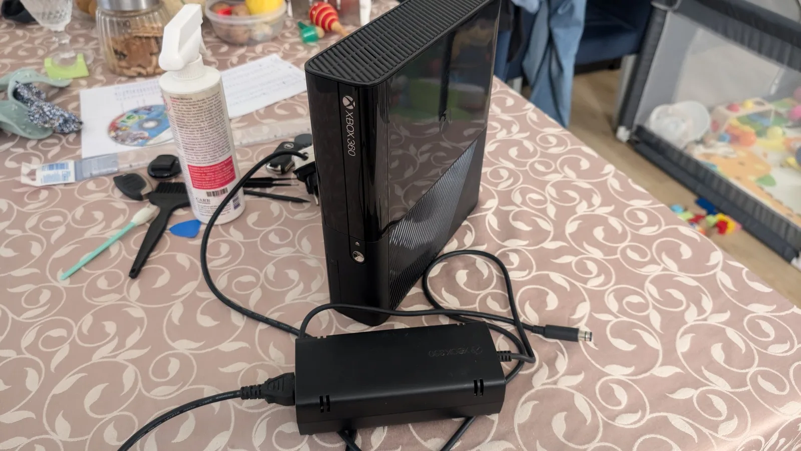Xbox 360 E console standing upright with its external power brick and cables on a patterned tablecloth, surrounded by cleaning supplies and pry tools.