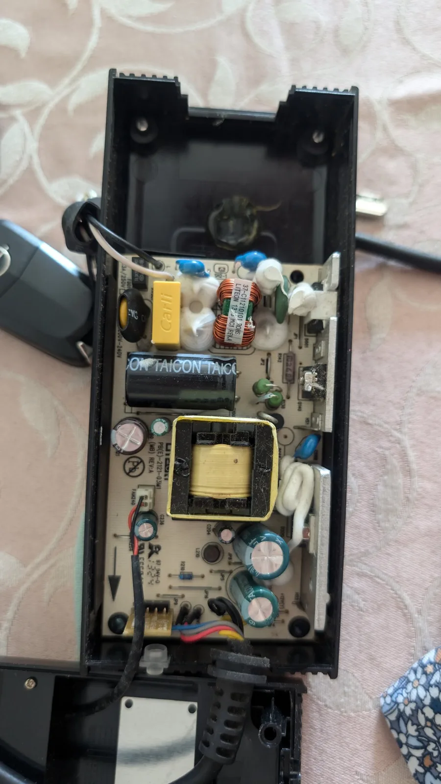 Opened Xbox 360 E power brick showing its LiteOn PSU board with the original bargain-bin capacitors before recapping, including a black Taicon electrolytic cap and a yellow box capacitor, plus the transformer and filter coil, on a patterned surface.