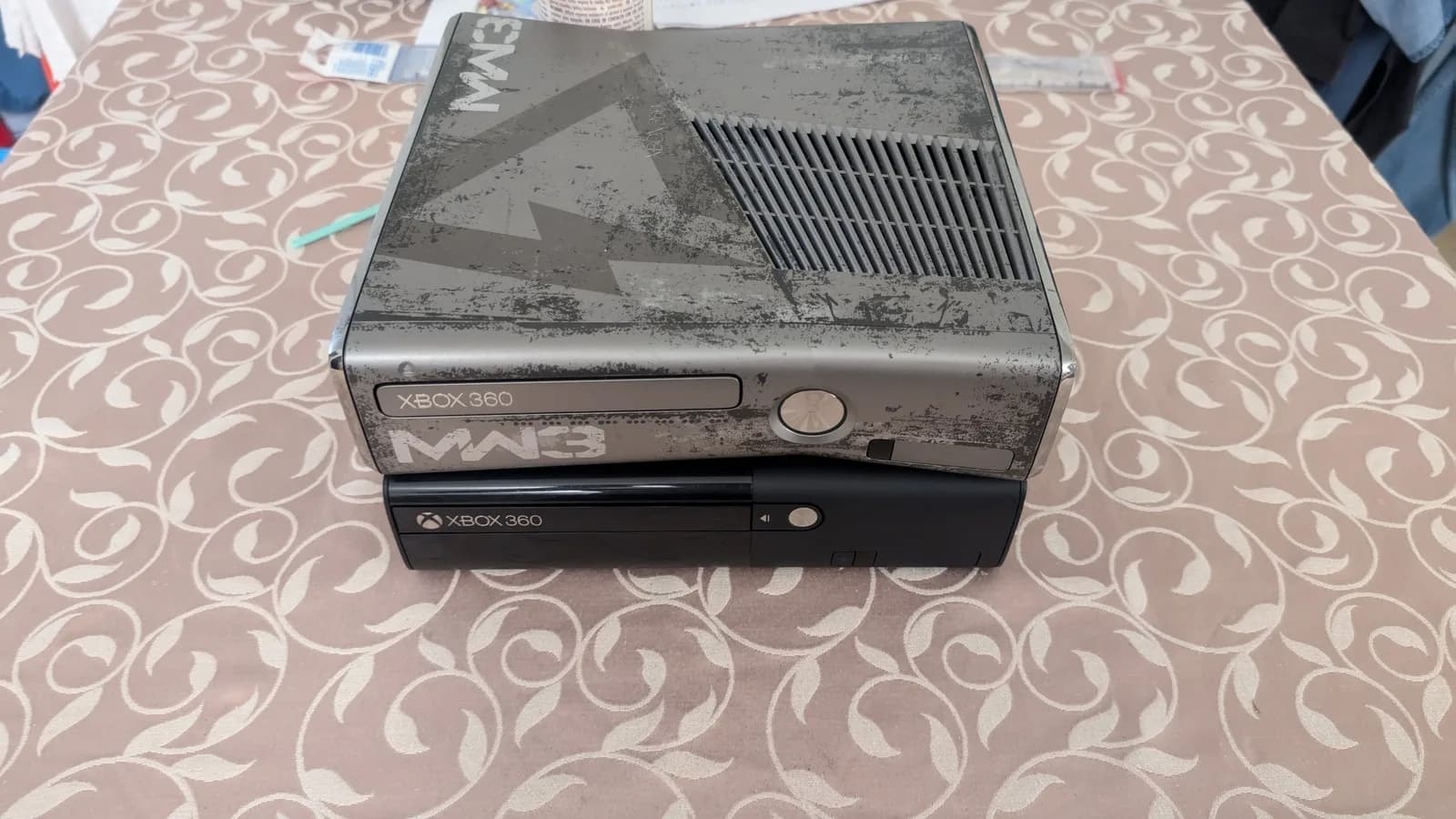Two Xbox 360 consoles stacked on patterned fabric: the dusty gray Modern Warfare 3 edition (a 360 S) on top, the glossy black 360 E below. Both show Xbox 360 branding.