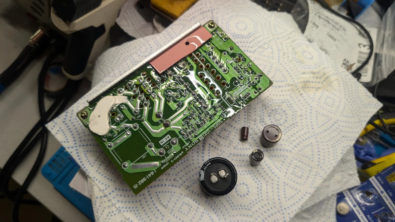 Reflowed solder side of the Sony PVM-9045QD PSU board (Sony 1-641-982-15) on a paper towel, glistening where it has been doused in isopropyl for cleaning, the removed electrolytic capacitors (including the large black ELNA main filter cap) set beside it during the recap.