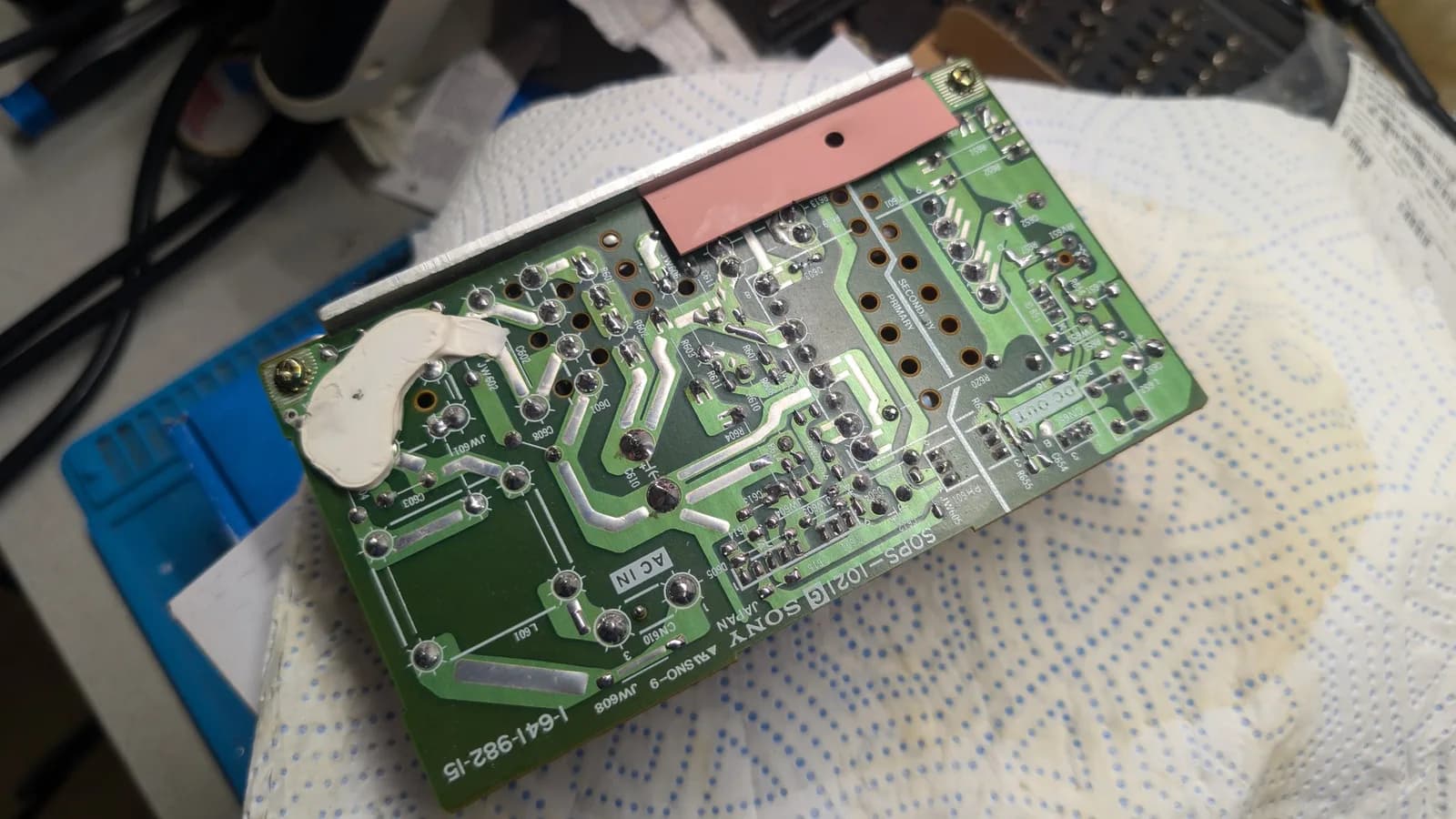 Solder side of the Sony PVM-9045QD PSU board (Sony 1-641-982-15) after reflow, the joints bright, with a pink rubbery pad and a bead of hardened white caulk near the board edge.