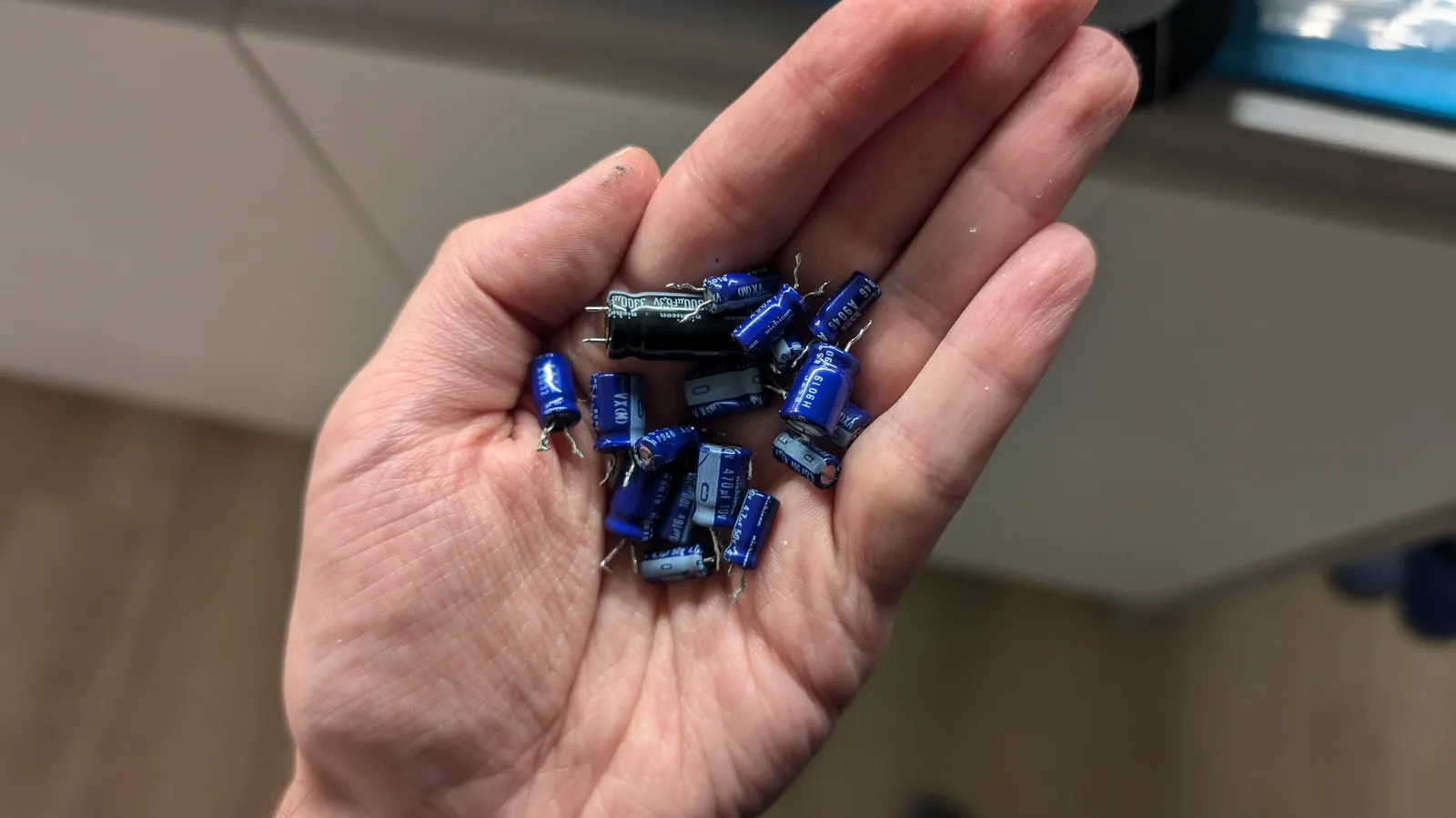 A hand holding the blue electrolytic capacitors removed during the partial mainboard recap, including one black capacitor among them.