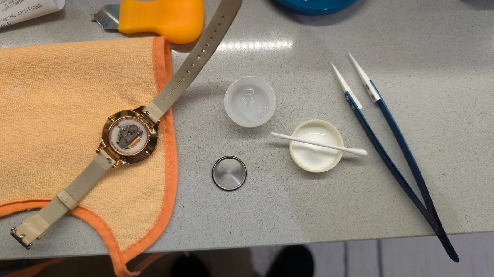 Watch repair workspace: disassembled watch with lubricated gasket, precision brushes, metal casing, palette with lubricant, ruler, and watch on orange fabric band.