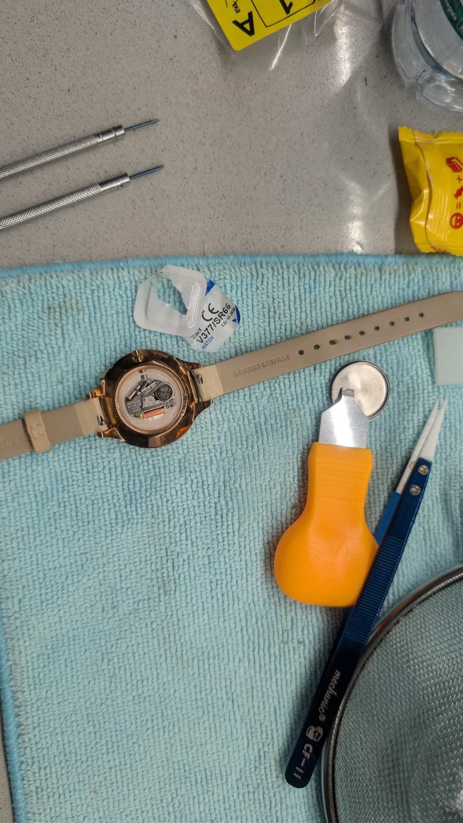Watchmaking tools laid out on light blue mat: open-faced watch with tan band, jeweler's screwdrivers, yellow bulb-shaped tool, precision picks, and repair supplies.