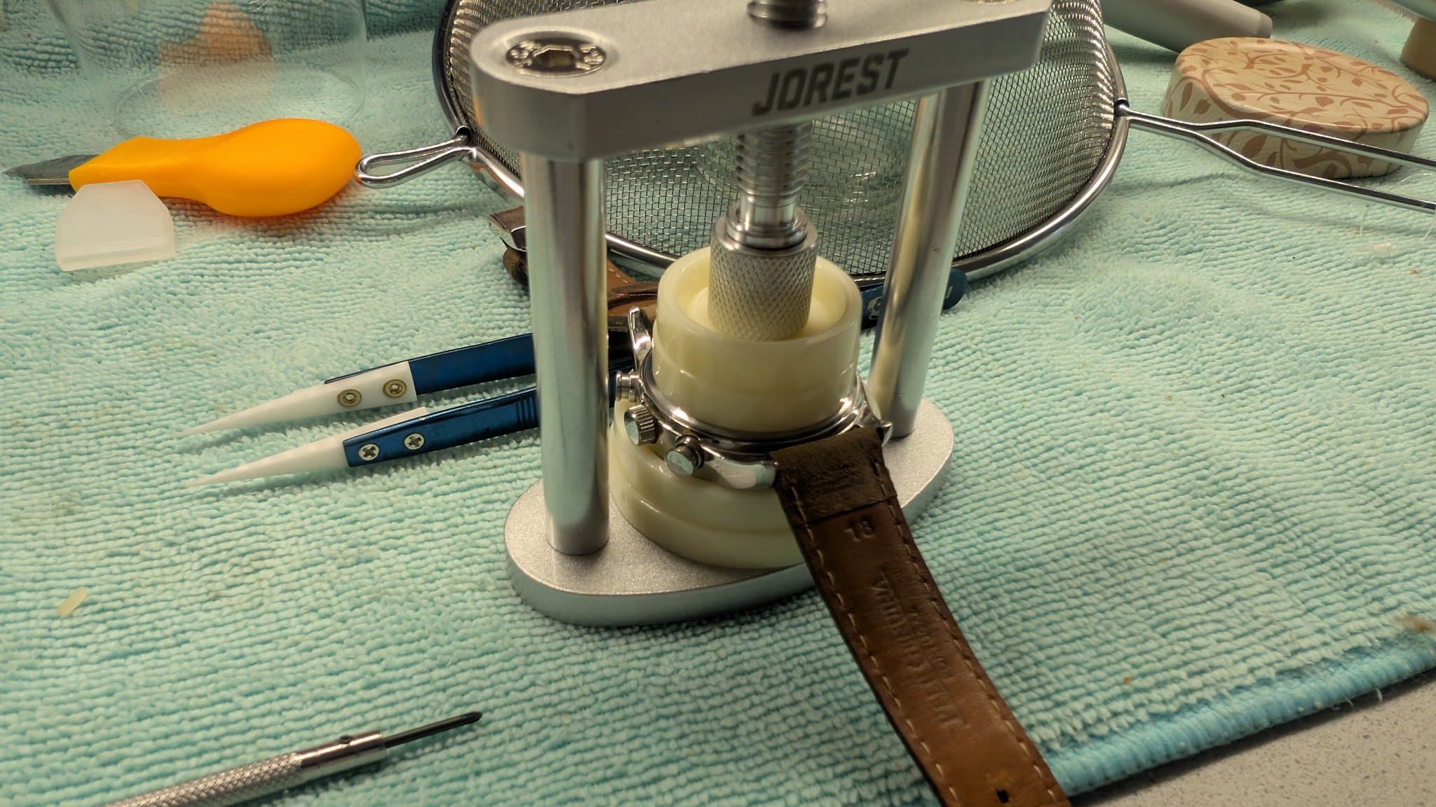 Jobrest watch case press tool with cream-colored platen, holding open watch case back; precision screwdrivers and orange battery case nearby on workmat.