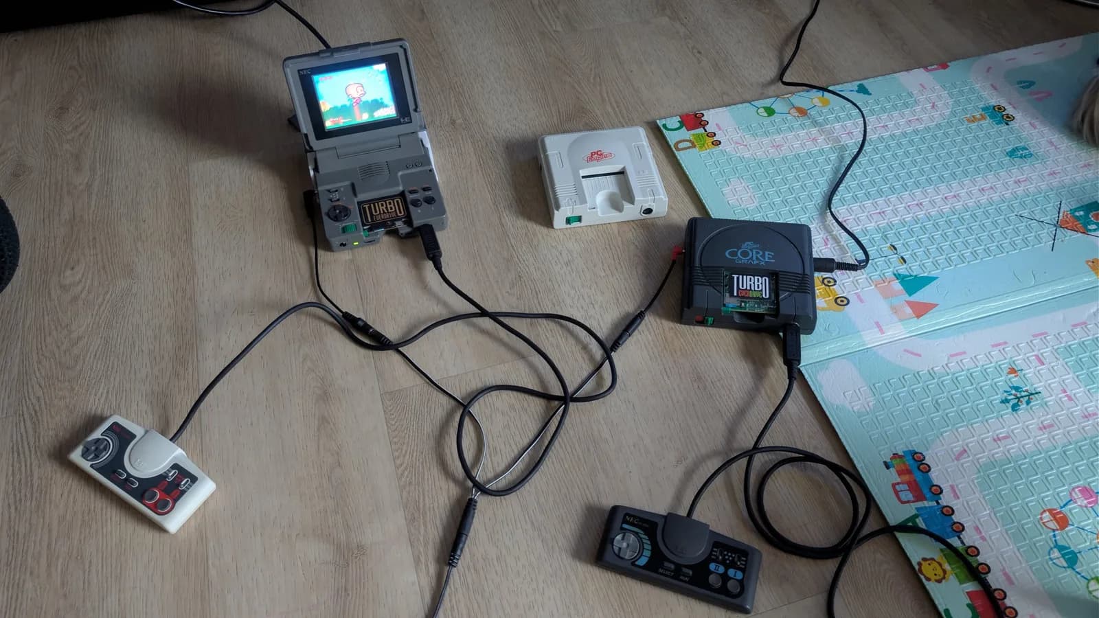 PC Engine hardware on a wooden floor for firmware testing: a PC Engine LT (the clamshell unit) with a Turbo Everdrive Pro, showing Bonk's Adventure on its built-in screen, and a grey PC Engine CoreGrafx with a Turbo Everdrive v2, plus controllers and tangled AV cables.