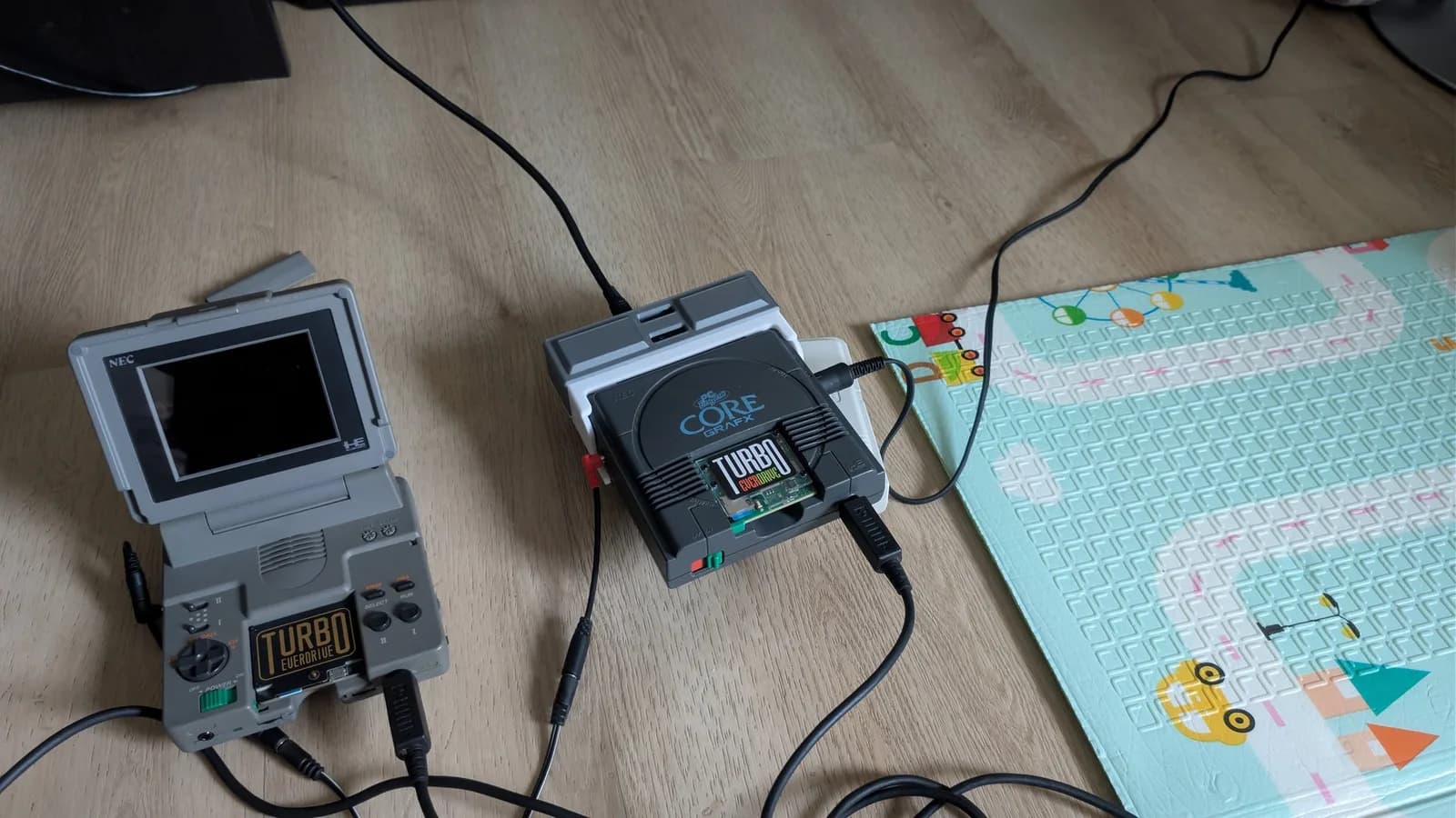 A PC Engine LT (the clamshell unit) with a Turbo Everdrive Pro and a grey PC Engine CoreGrafx with a Turbo Everdrive v2, on a wooden floor beside a child's foam play mat, AV cables connected for firmware testing.