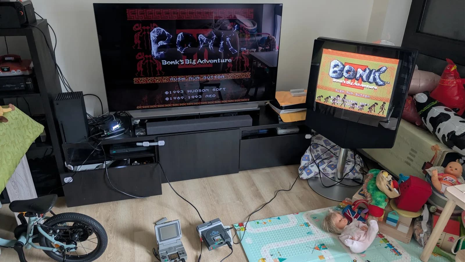 Bonk's Big Adventure title screen (Hudson Soft, 1993) on two displays in a living room, the same game on each to compare the OSSC's custom firmware (on the flat-panel TV) against the CRT's direct output; a PC Engine LT and a grey CoreGrafx on the floor among toys and a child's play mat.