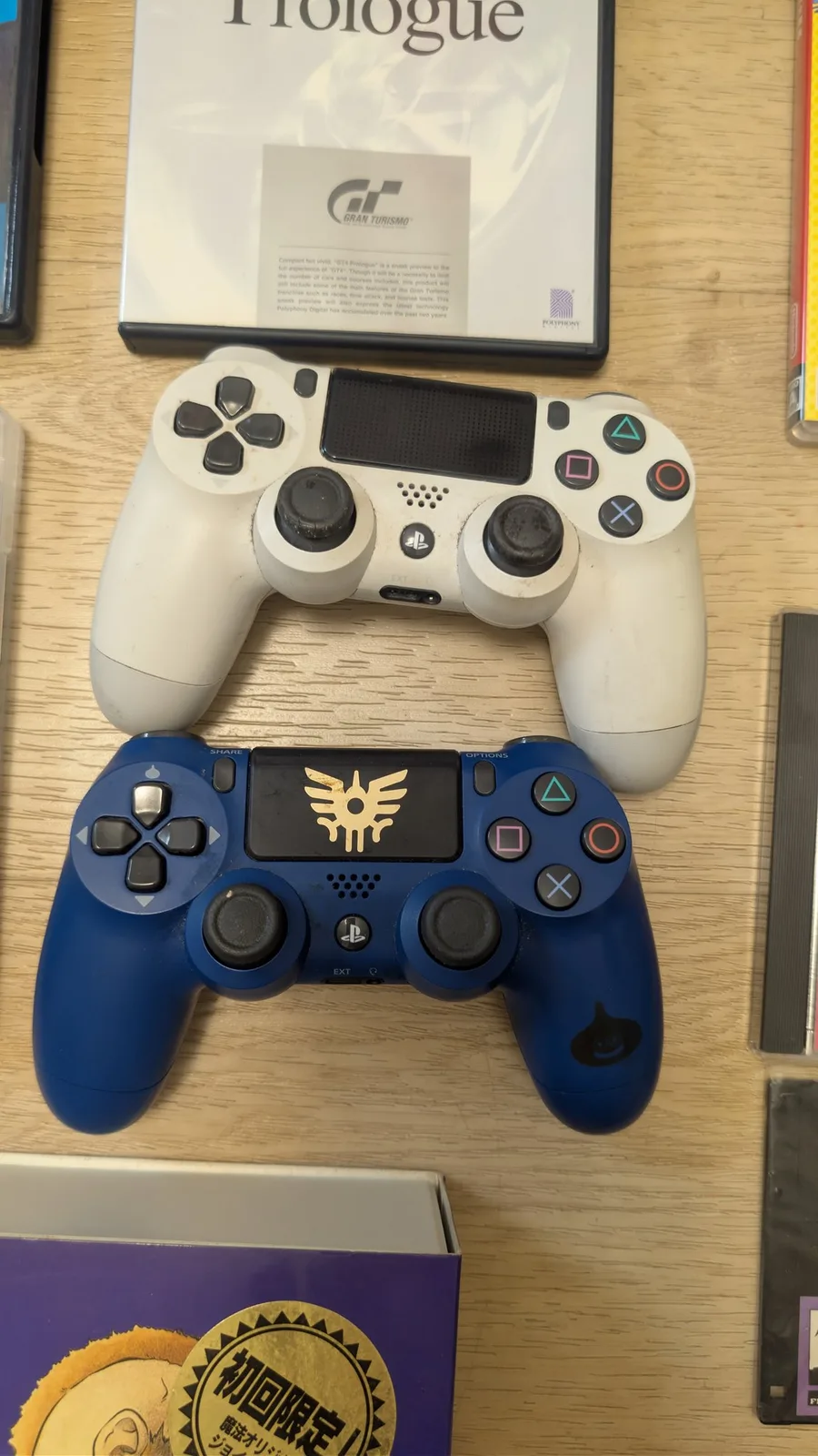 Two PS4 DualShock 4 controllers on a wooden surface before refurbishment: a grimy white controller on top with dirty, worn thumbsticks, and a dark blue Dragon Quest special-edition controller below with a gold winged crest on the touchpad and a gold slime character on the lower grip.