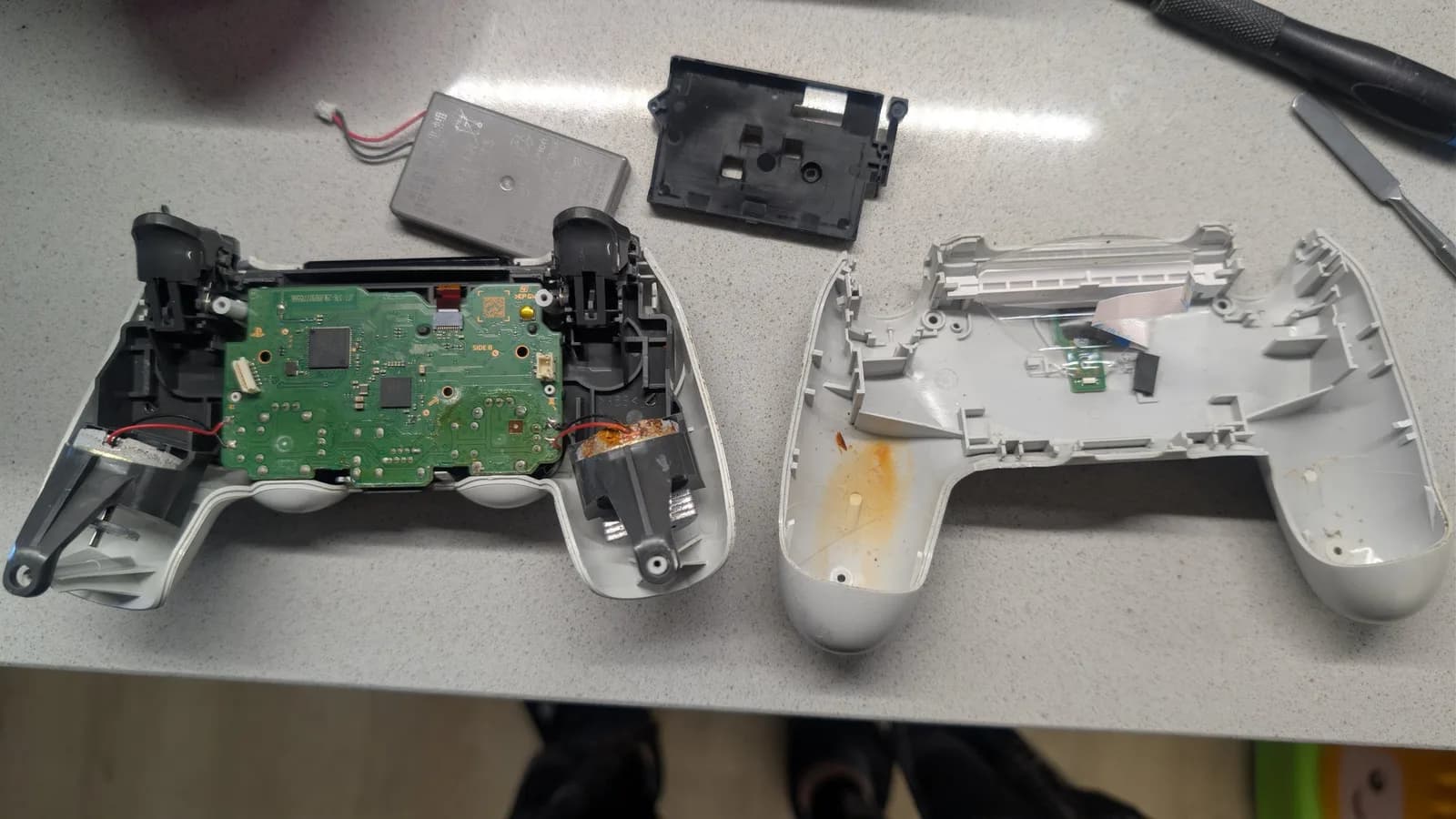Disassembled white PS4 controller: the front shell at left still holds the green mainboard, with a rumble motor at the board's right edge showing orange rust; the silver lithium-ion battery and its black holder sit above, and the back shell half at right has a matching orange rust stain inside. Screwdrivers at top right.