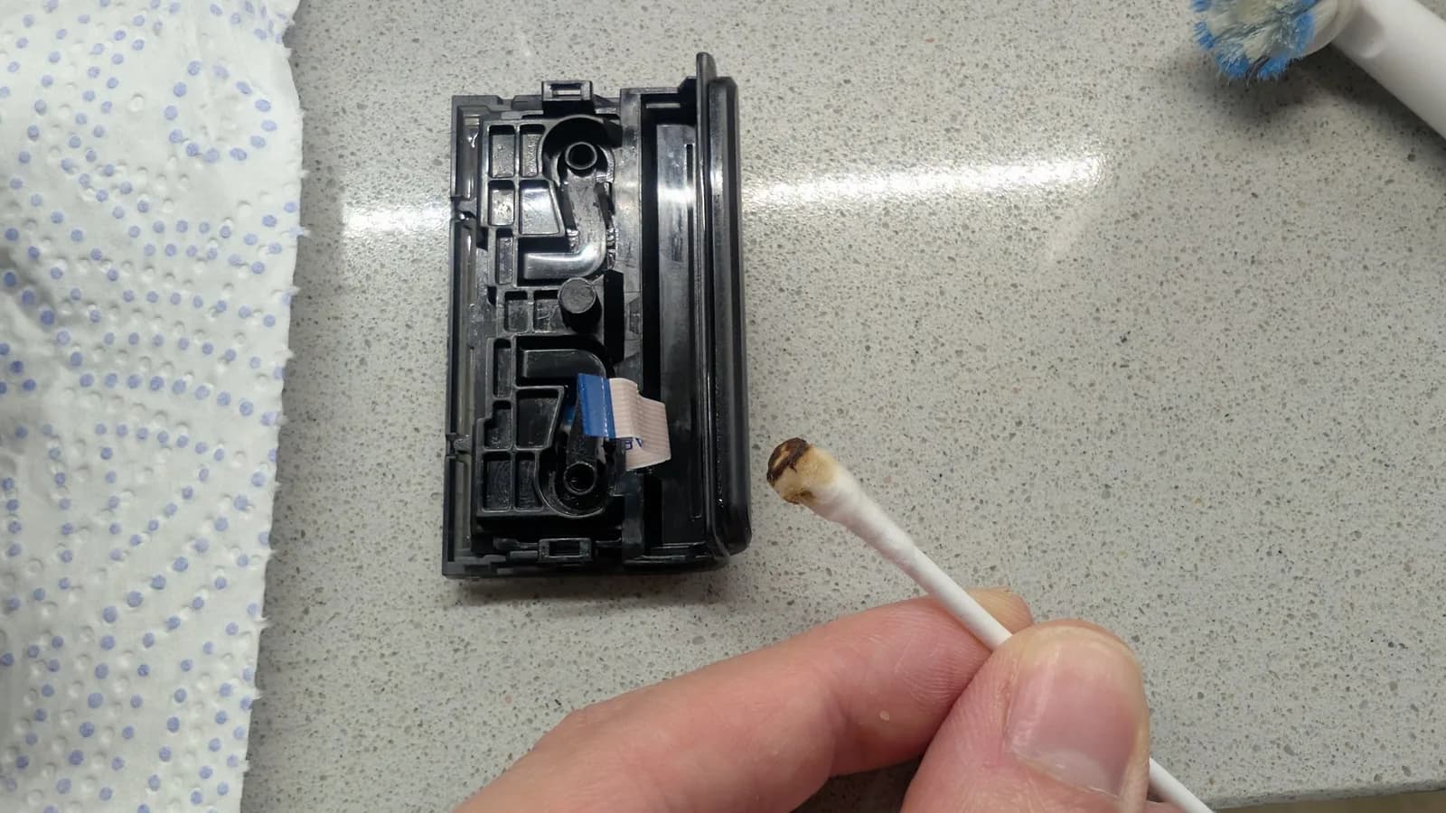 The controller's black touchpad module with its white ribbon cable (blue tab), being cleaned with a cotton swab whose tip is now brown with grime; a brush head at top right.
