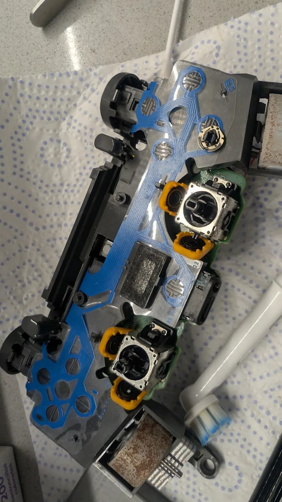Disassembled PS4 controller mainboard on paper towel: its membrane flex (blue tracks, black carbon button contacts) over the grey plastic frame, two analog stick modules with orange potentiometer casings (grimy and worn) at centre, and the rumble motors in their housings showing heavy orange rust at top right and bottom. Just above the topmost thumbstick, the wiper that normally sits inside the potentiometer casing (it rotates as the stick moves) rests on the membrane. A toothbrush head alongside.