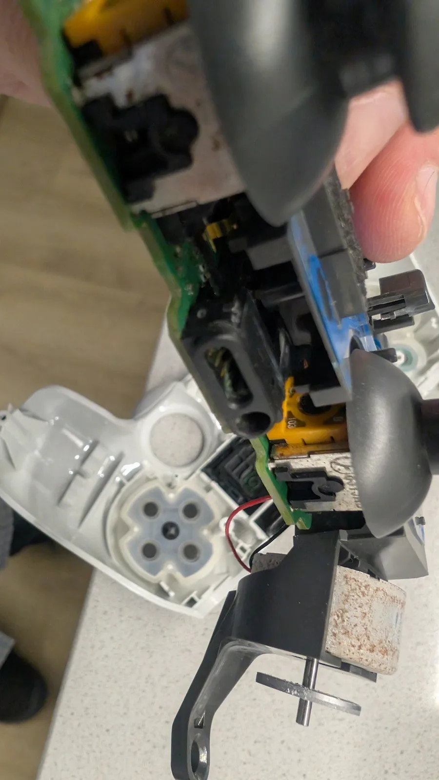 Close-up of a white PS4 controller's mainboard held up during cleaning, centred on the black EXT extension port with green corrosion on the pins next to the round 3.5mm headset jack; green PCB, an orange analog-stick potentiometer and the white shell's cross-shaped D-pad membrane behind.
