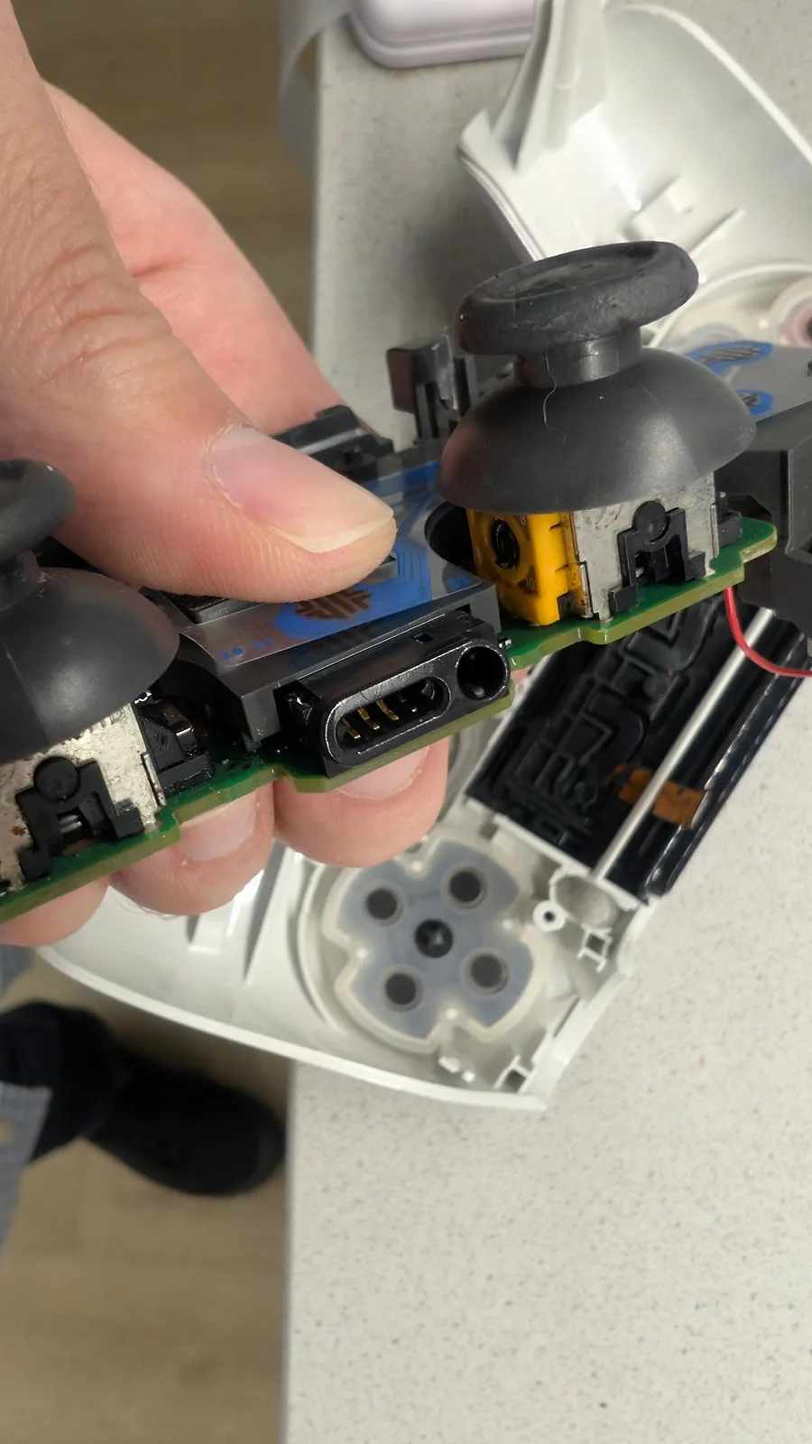 Close-up of the same white PS4 controller board held in the fingers after cleaning: the black EXT extension port's pins now shining and corrosion-free next to the round 3.5mm headset jack in the foreground, the two black-capped analog sticks with their orange potentiometers on either side of the port, and the white shell's D-pad membrane behind.