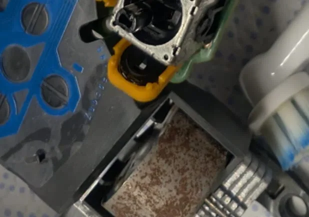Close-up of the drift repair: the analog stick's orange potentiometer casing split away from its metal frame so the carbon track inside can be cleaned with a small white and blue brush; a face-button membrane with blue tracks at left.