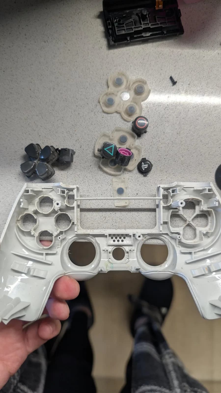 A hand holds the white front faceplate of a disassembled PS4 controller; laid out above it are the grimy parts removed from it: the rubber button membranes, the D-pad, the four face buttons and the PS button, and the touchpad at top.